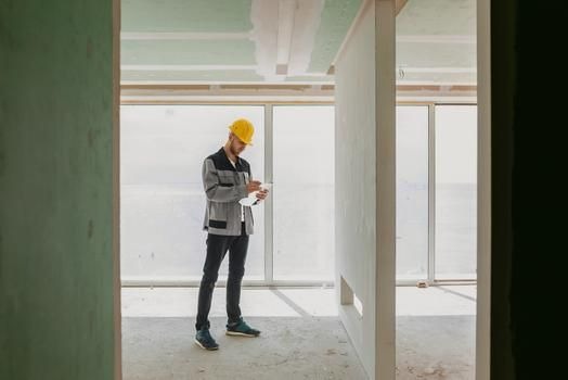 Inspektor w kasku, z dokumentami, dokonuje odbioru mieszkania w stanie deweloperskim. Widoczne surowe ściany, okna i elementy konstrukcyjne.