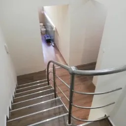 Klatka schodowa z drewnopodobnymi płytkami i metalową balustradą, widok z g&oacute;ry, w tle widoczne pomieszczenie z białymi ścianami i podłogą z paneli.