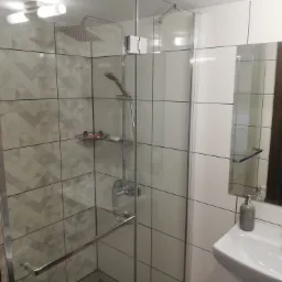 Nowoczesna kabina prysznicowa z deszczownicą i szklanymi drzwiami, widoczna umywalka z szarą pompką na mydło i lustro z odbiciem ciemnego drewna drzwi.