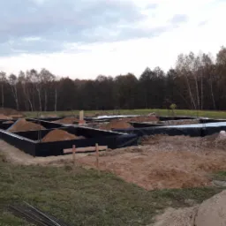 Przygotowanie terenu pod budowę: wykopane fundamenty z czarną izolacją, częściowo zasypane piaskiem, na działce z widokiem na las pod pochmurnym niebem.