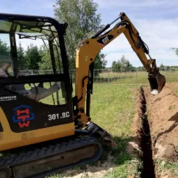 Żółta mini koparka CAT 301.8C wykonuje wąski wykop w ziemi na trawiastym terenie w słoneczny dzień, widoczne drzewa w tle.