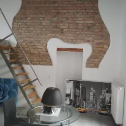 Wnętrze z ceglaną ścianą o nieregularnym kształcie, schodami na antresolę i okrągłym szklanym stołem z szarymi krzesłami.