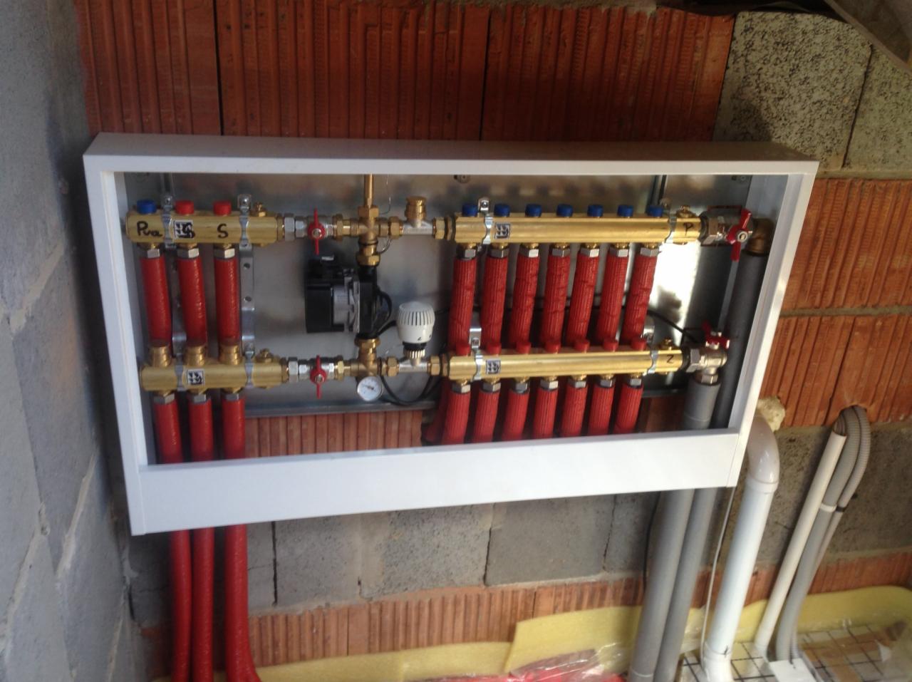 Rozdzielacz ogrzewania podłogowego w białej szafce, widoczne czerwone pętle, złote rury, pompa obiegowa i manometr, zamontowany na ścianie z cegły
