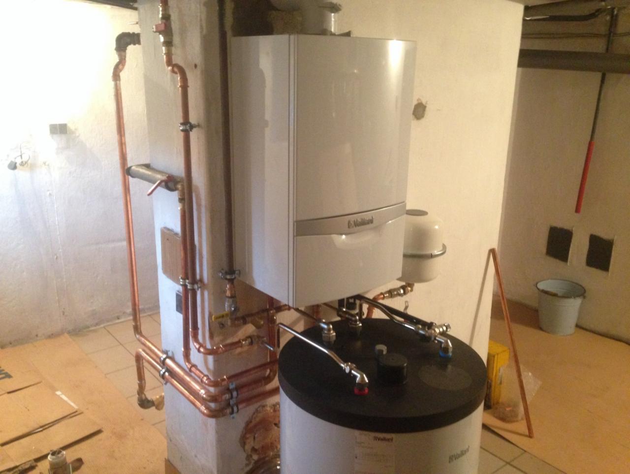 Nowoczesny, biały piec gazowy Vaillant zamontowany na ścianie, z widoczną instalacją miedzianą i zasobnikiem, w pomieszczeniu z płytkami podłogowymi.