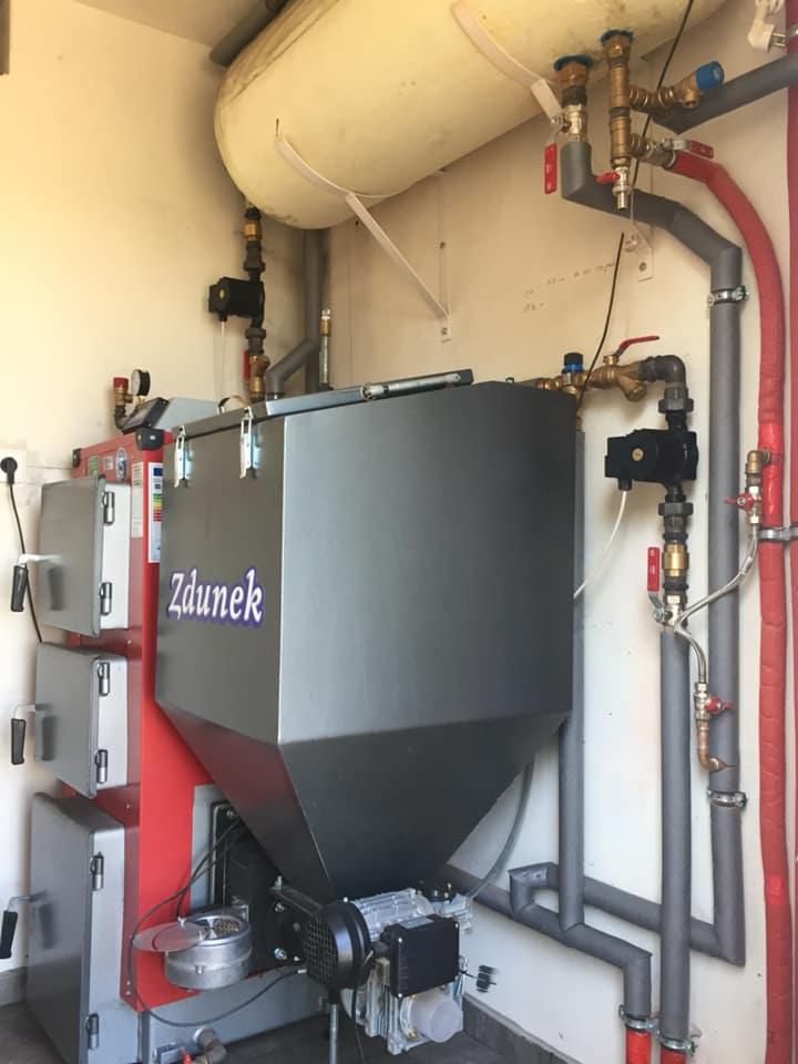 Zamontowany piec na paliwo stałe z zasobnikiem, widoczne rury instalacji CO i CWU oraz naczynie wzbiorcze, nazwa producenta 'Zdunek' na obudowie.