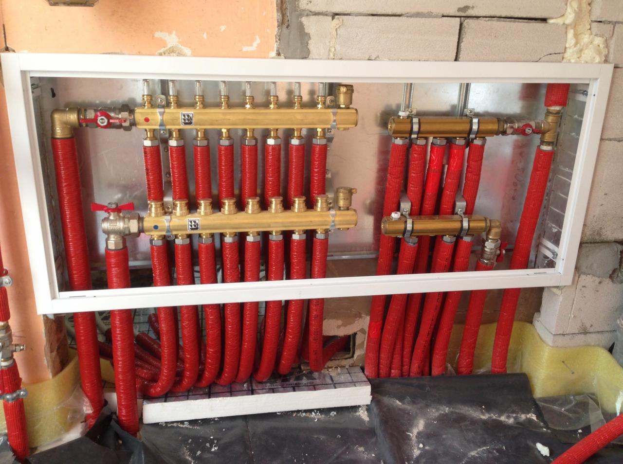 Rozdzielacz ogrzewania podłogowego z czerwonymi rurami w szafce z białym obramowaniem, widoczne elementy montażowe i fragment surowej ściany w tle.
