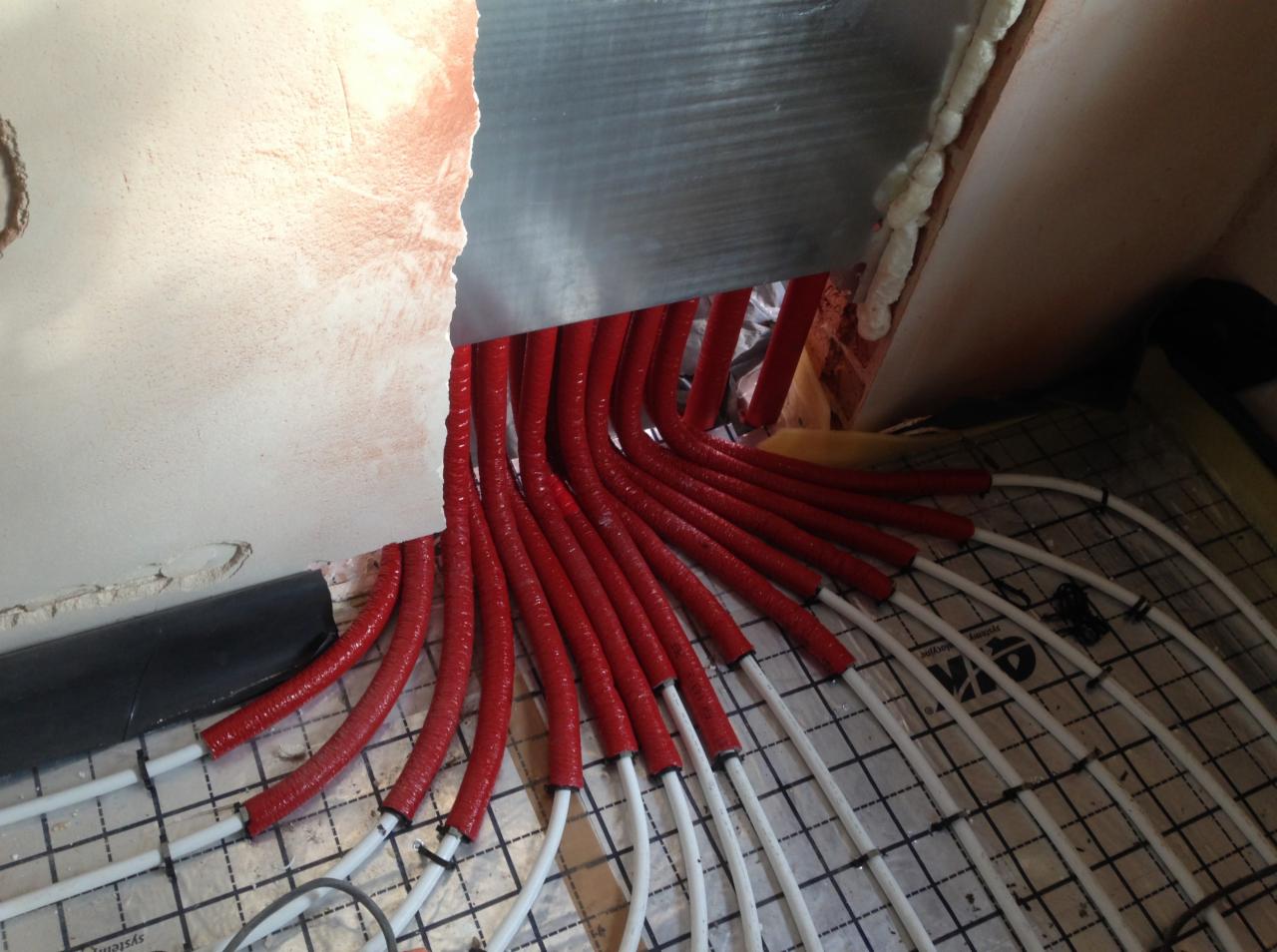 Czerwone rury grzewcze podłogowe wychodzące ze ściany, częściowo zaizolowane, układane na siatce montażowej w trakcie instalacji.