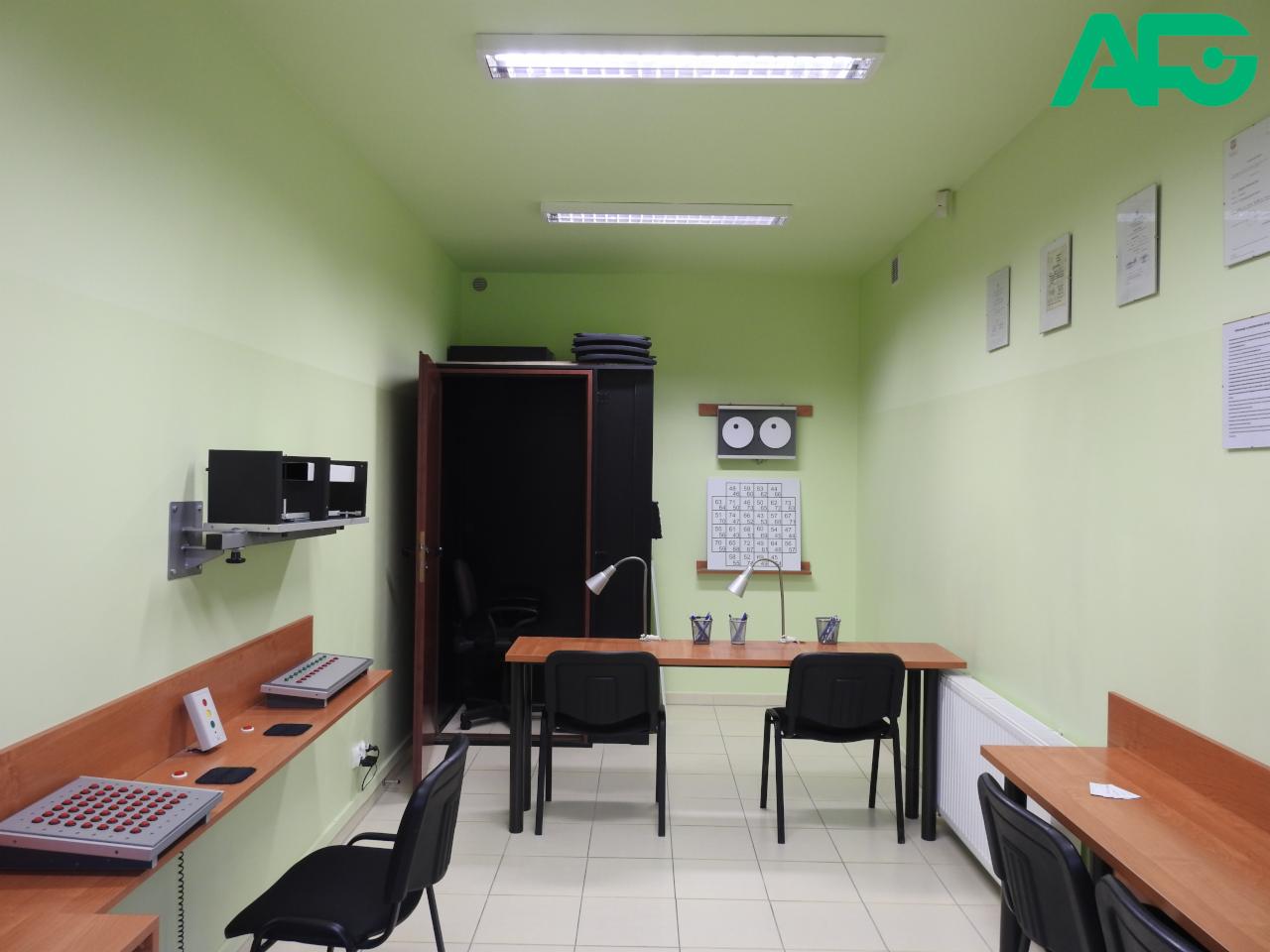 Gabinet z wyposażeniem do badań słuchu: audiometr na ścianie, kabina dźwiękoszczelna, biurka z lampkami, kalendarz i dokumenty na ścianie.