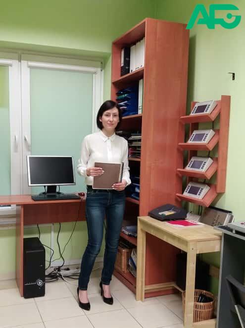 Psycholog w jasnym gabinecie, stojąca obok biurka z komputerem, trzymająca notatnik.