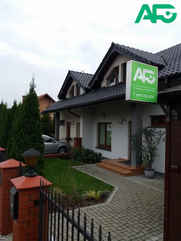 Budynek poradni psychologicznej z zielonym szyldem i numerem telefonu, otoczony zadbanym ogrodem i ogrodzeniem.