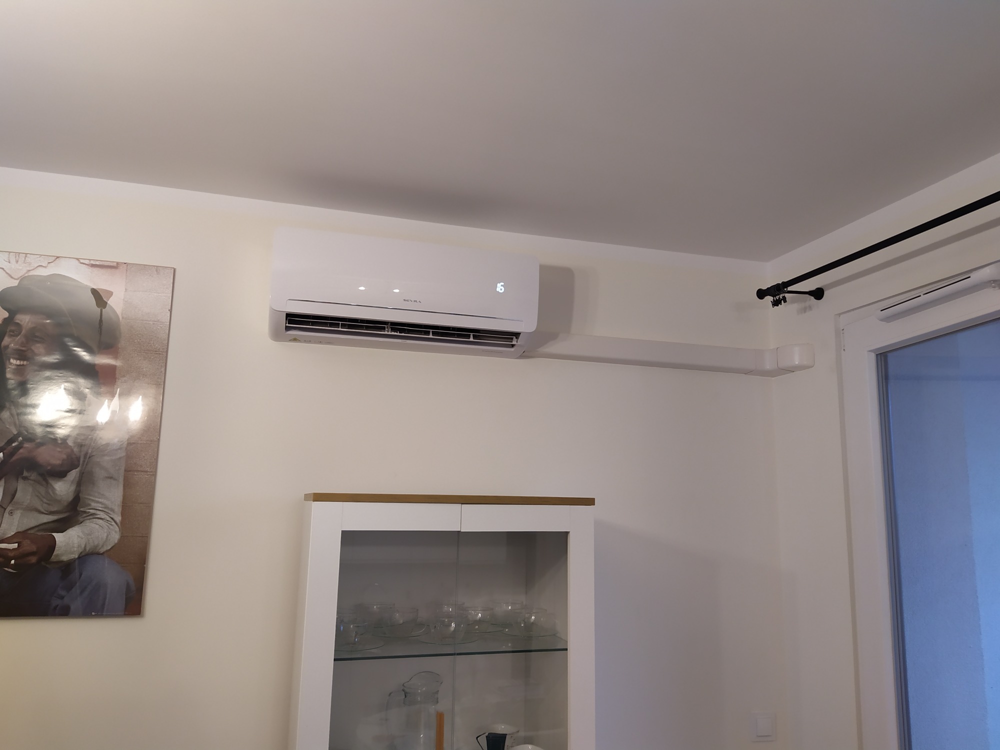 Biały klimatyzator ścienny marki Sevra z wyświetlaczem temperatury '16' zamontowany w pomieszczeniu mieszkalnym, z widoczną instalacją elektryczną w białej listwie i obrazem na ścianie.