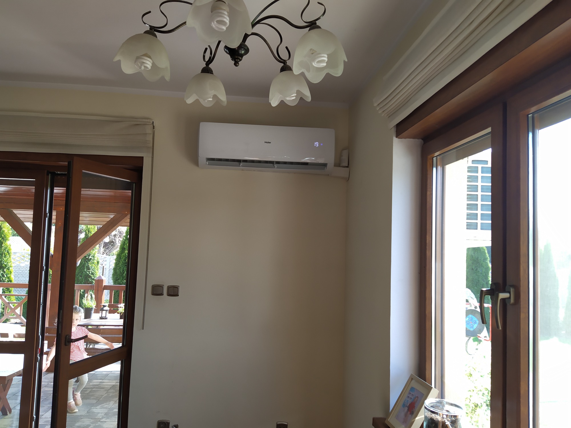 Biały klimatyzator Haier zamontowany na ścianie w jasnym pokoju, widoczny wyświetlacz z temperaturą 19 stopni, pod spodem dekoracyjny żyrandol z kwiatowymi kloszami, w tle otwarte drzwi balkonowe...