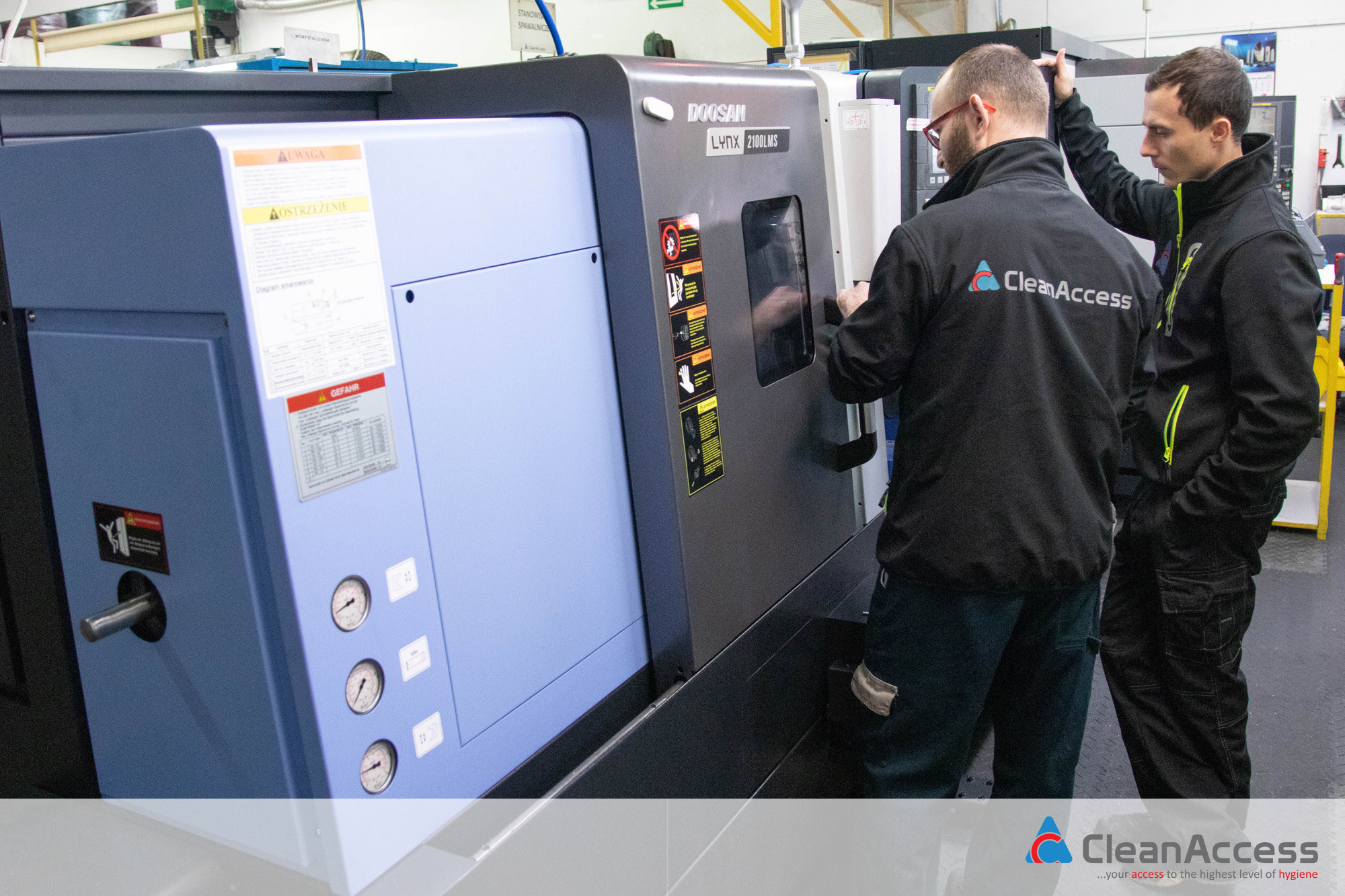 Dwóch mężczyzn w firmowych kurtkach CleanAccess obsługuje nowoczesną tokarkę CNC Doosan Lynx 2100LMS w hali produkcyjnej.