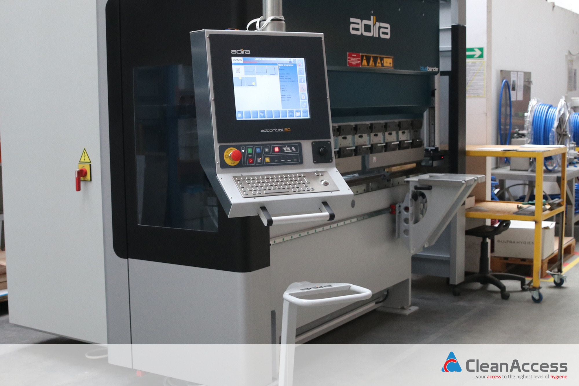 Nowoczesna prasa krawędziowa CNC Adira z panelem sterowania adcontrol 60, widoczny ekran z listą programów i klawiatura, w tle elementy wyposażenia hali produkcyjnej.