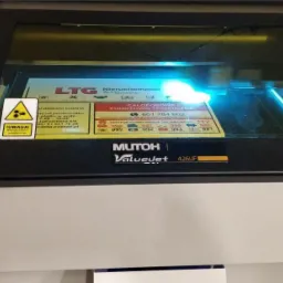 Drukarka UV Mutoh ValueJet 426UF w trakcie druku z niebieskim światłem, widoczna etykieta z ostrzeżeniem o promieniowaniu.