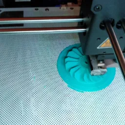 Druk 3D turbiny wentylatora z tworzywa w kolorze turkusowym na perforowanym stole roboczym drukarki 3D, widoczne dysza drukująca i elementy mechaniczne drukarki.