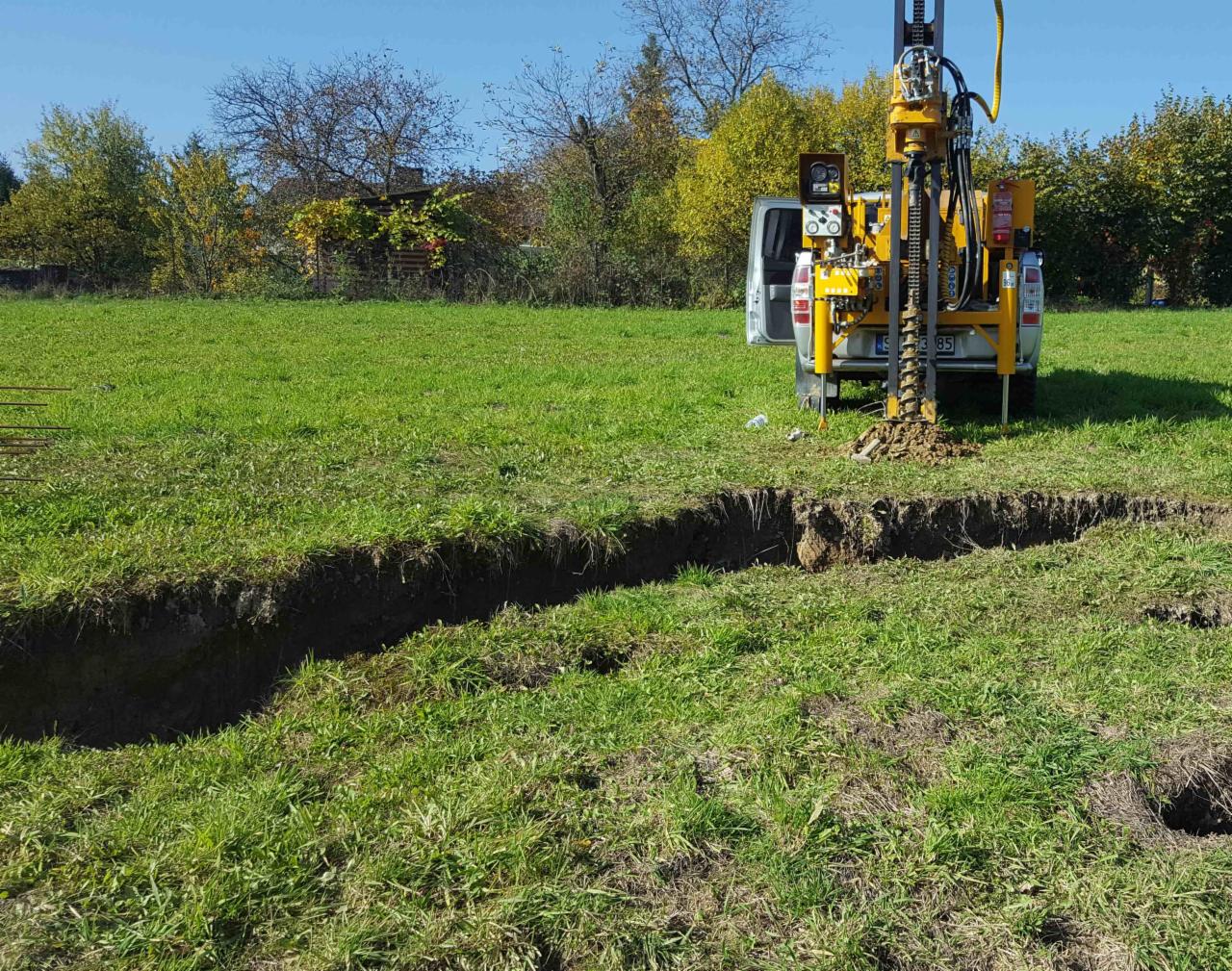 Żółty wiertniczy pojazd geologiczny wiercący w zielonym polu, z widoczną otwartą tylną klapą pojazdu i wykopem w ziemi.