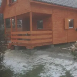 Drewniany dom z bali z zadaszonym gankiem, szara blachodachówka, widoczne fundamenty z betonowych bloczków, trawa z resztkami śniegu.