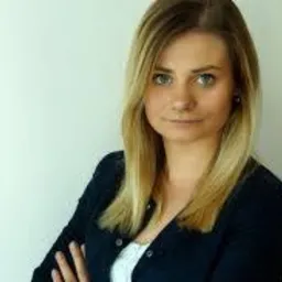 Portret młodej kobiety z blond włosami, ubrana w ciemną marynarkę i białą bluzkę, z założonymi rękami, patrząca prosto w obiektyw na jasnym tle.