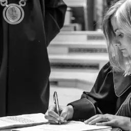 Kobieta prawnik w todze podpisuje dokumenty, w tle fragment osoby w uroczystym stroju z łańcuchem.