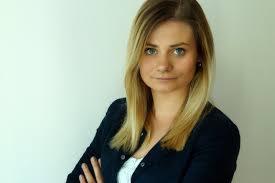 Portret młodej kobiety z blond włosami i niebieskimi oczami, ubrana w ciemną marynarkę i białą bluzkę, pozująca na jasnym tle.