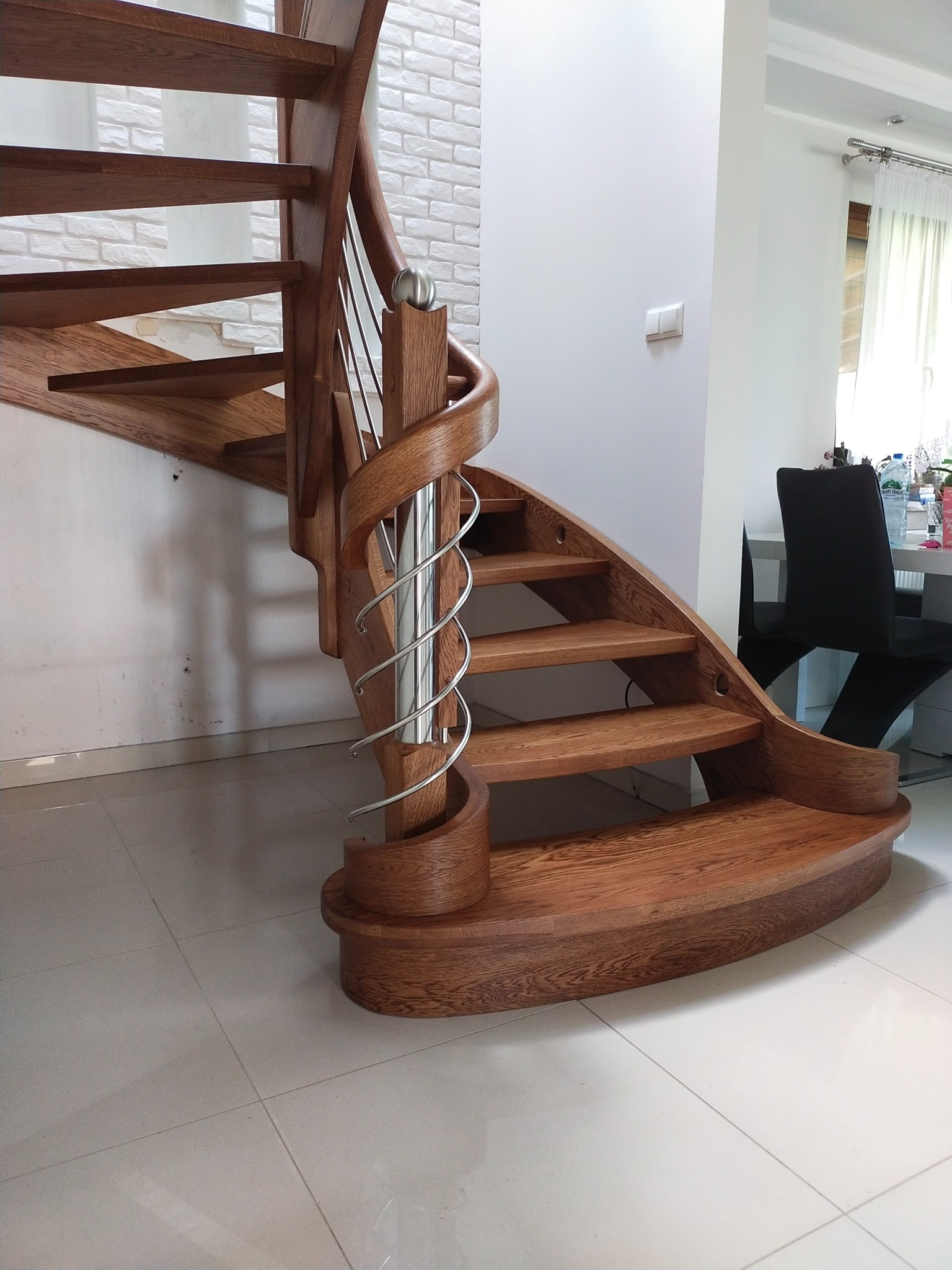 Kręte, dębowe schody z metalową balustradą, elegancko wkomponowane w jasne wnętrze. Widoczne detale wykończenia i naturalna barwa drewna.
