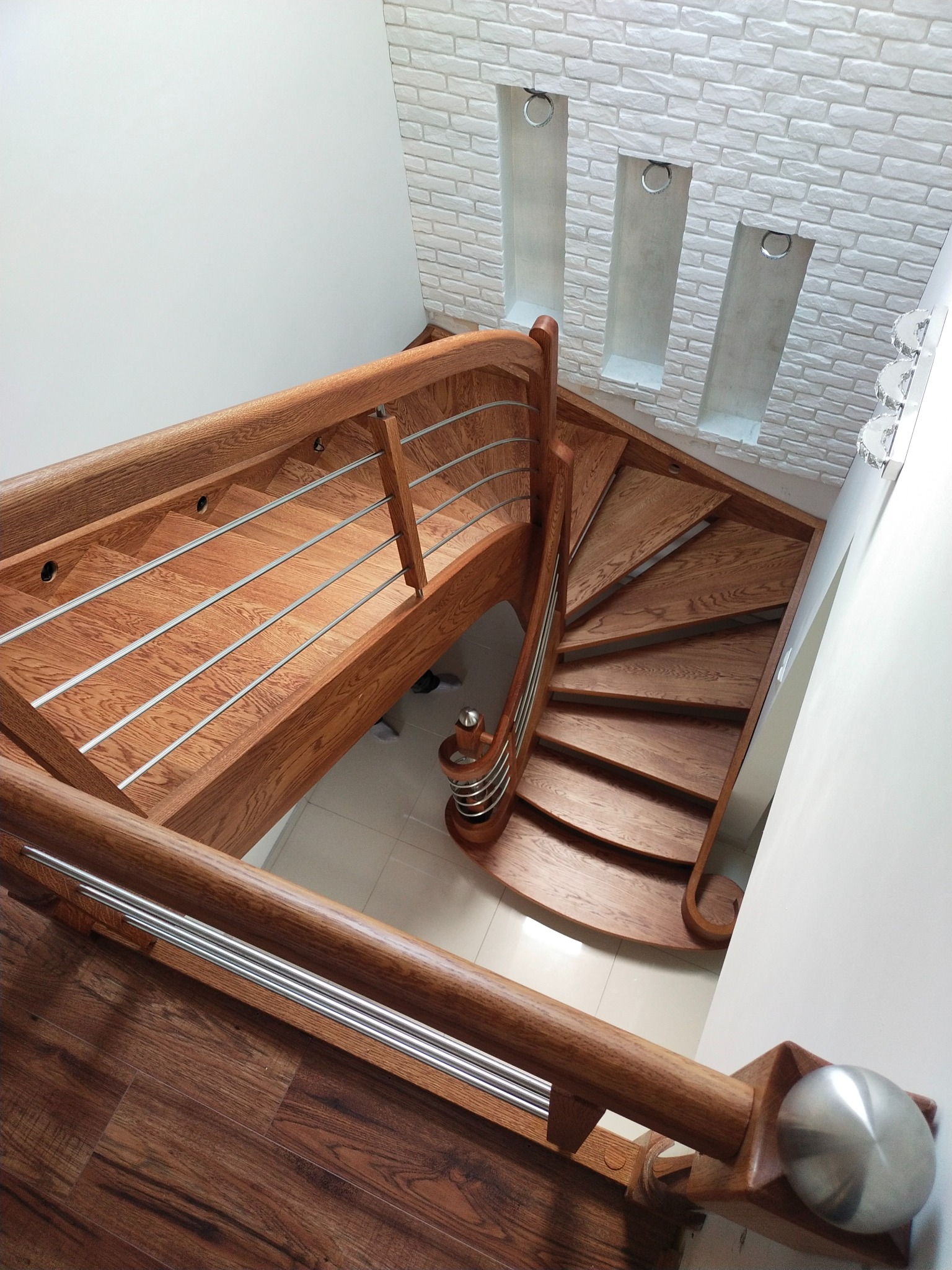 Kątowe ujęcie dębowych schodów z metalowymi balustradami, z widoczną spiralną konstrukcją i dekoracyjną ścianą z cegły w tle. Eleganckie wykończenie.