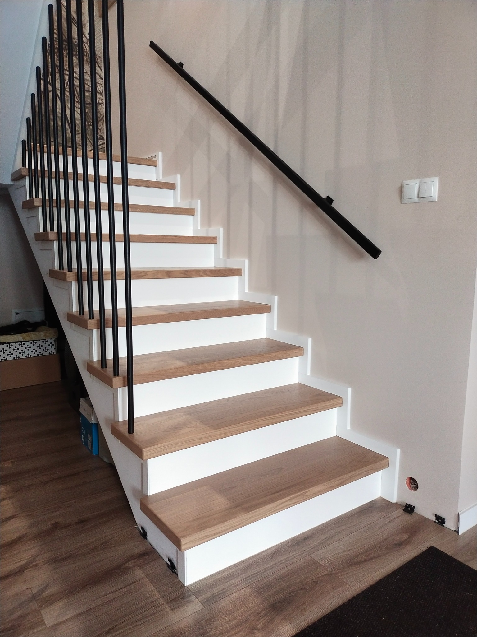 Nowoczesne schody z drewnianymi stopniami i czarną, metalową balustradą. Jasne ściany i podłoga imitująca drewno. Widoczne detale wykończenia.