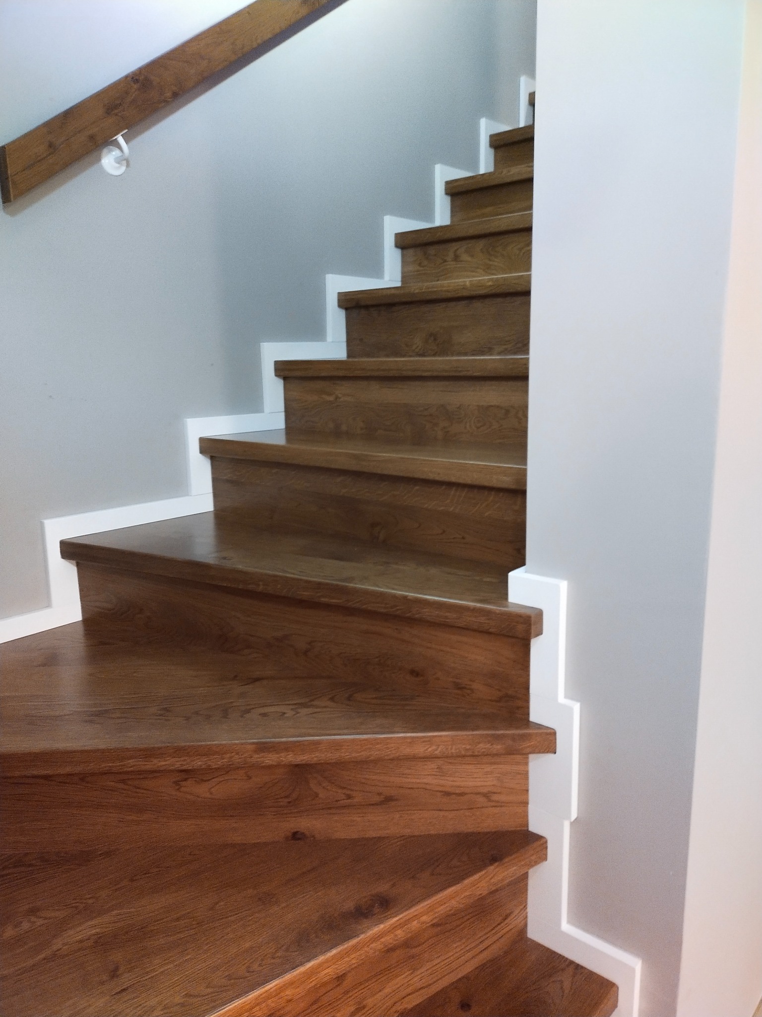 Nowoczesne, drewniane schody z białymi podstopnicami i drewnianą poręczą, oświetlone naturalnym światłem, widok z dołu na pierwsze stopnie.