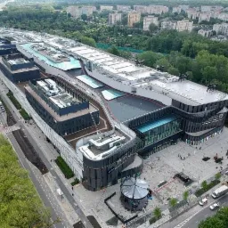 Szeroki kadr z lotu ptaka na nowoczesne centrum handlowe z przeszklonymi wejściami i częściowo odsłoniętym dachem, otoczone zielenią i zabudową miejską.