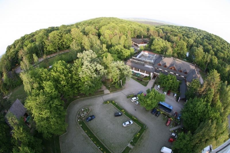 Szeroki kadr z lotu ptaka na hotel otoczony lasem, z widocznym parkingiem i wejściem.