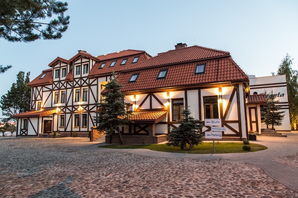 Elewacja budynku w stylu pruskim z oknami i oświetleniem zewnętrznym, widoczny bruk na podjeździe i tabliczki informacyjne.