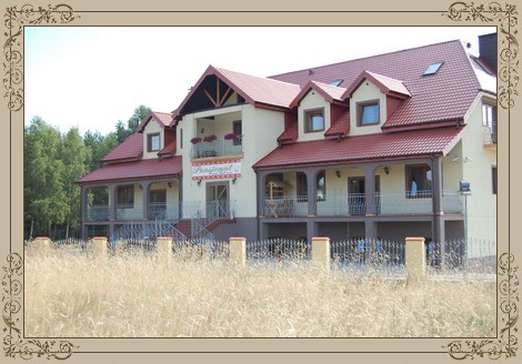 Elewacja budynku pensjonatu z czerwonym dachem, balustradami balkonowymi i ozdobnym ogrodzeniem, widoczna zza pola wyschniętej trawy.