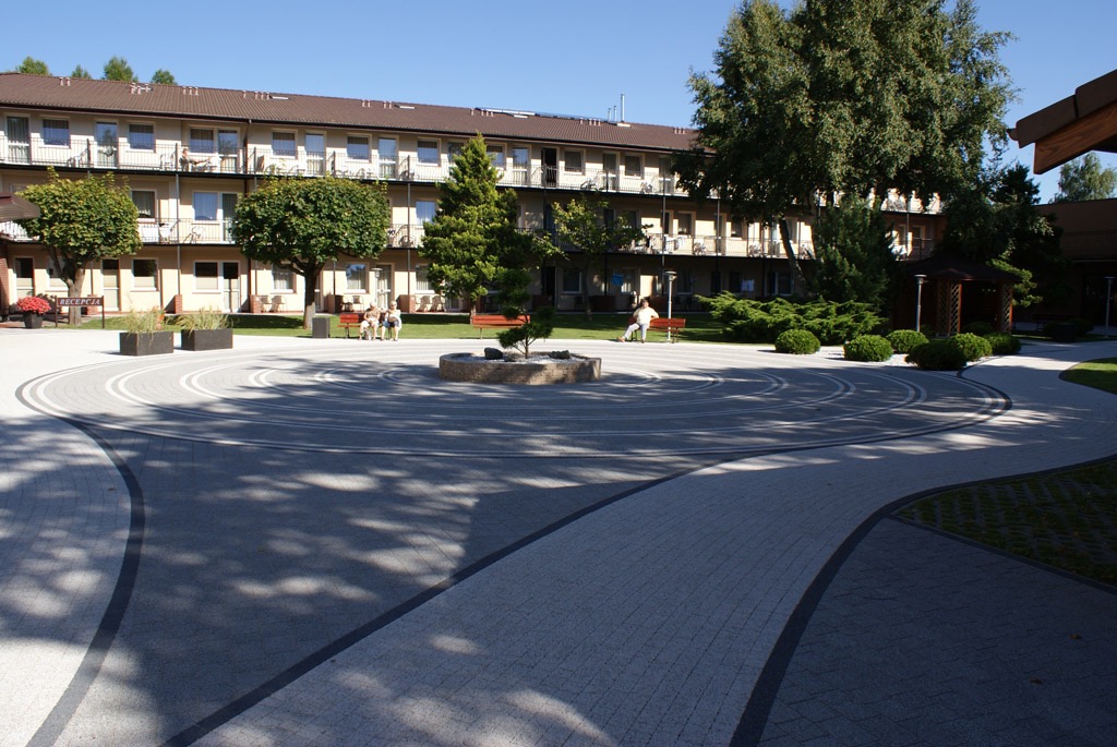 Dziedziniec z kostki brukowej ułożonej w koncentryczne okręgi, z małą fontanną pośrodku, otoczony zielenią i budynkiem z balkonami.