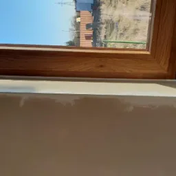 Drewniana rama okna z widoczną taśmą uszczelniającą 'WINDOWS TAPE INSIDE', częściowo wykończona ściana z widocznymi śladami po szpachlowaniu.