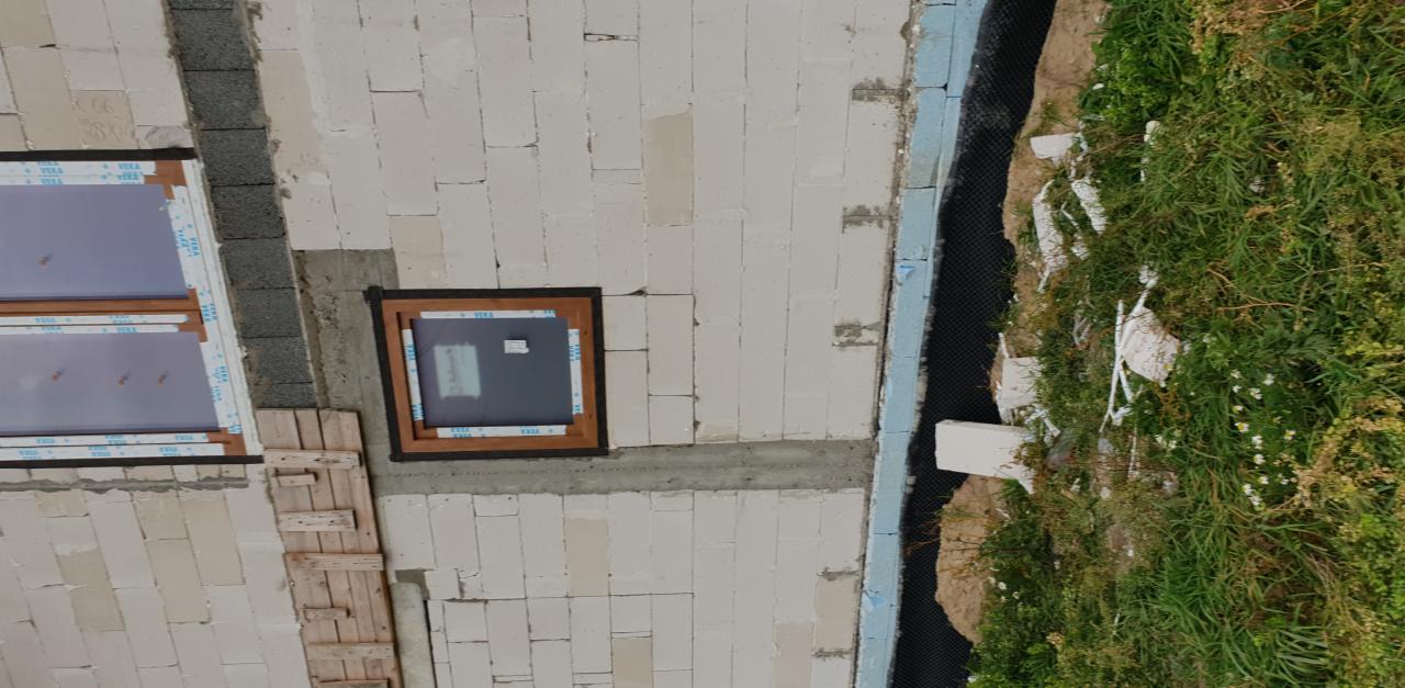 Widok z góry na budowę domu z widocznymi wstawionymi oknami i przygotowanym otworem na kolejne okno, zabezpieczonym drewnianą konstrukcją, oraz izolacją fundamentów od strony porośniętej trawą.