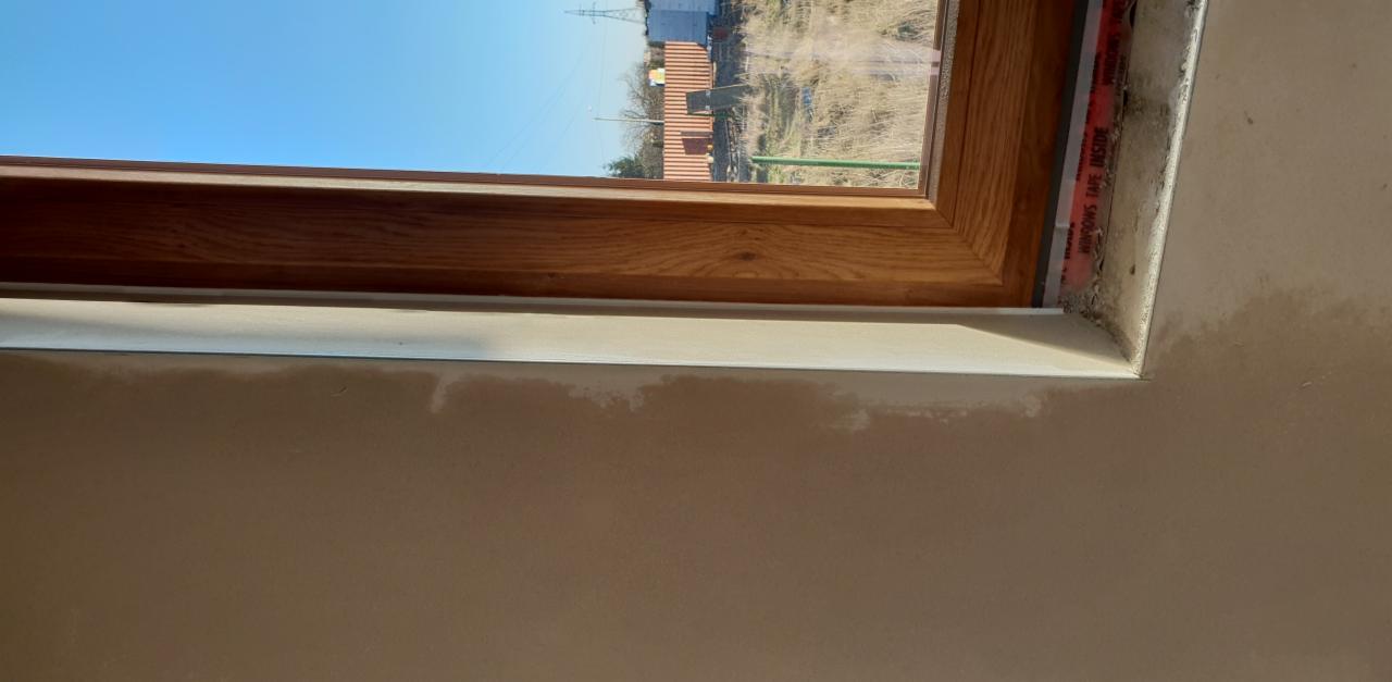 Drewniana rama okna z widoczną taśmą uszczelniającą 'WINDOWS TAPE INSIDE', częściowo wykończona ściana z widocznymi śladami po szpachlowaniu.
