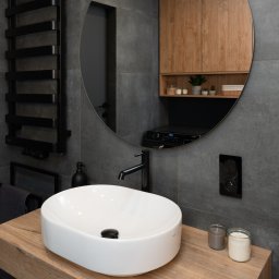 Triangle Studio - Nowoczesna łazienka z owalną umywalką nablatową KOLO na drewnianym blacie, czarną armaturą i grzejnikiem drabinkowym na tle szarych płytek.