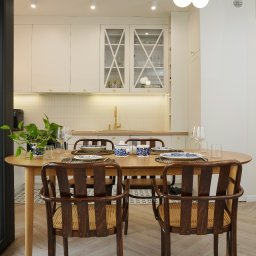 Triangle Studio - Jasna kuchnia z drewnianym stołem i krzesłami w stylu retro. Na stole zastawa w niebieskie wzory. W tle białe szafki z przeszklonymi frontami i złota armatura.