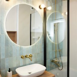 Triangle Studio - Nowoczesna łazienka z owalną umywalką, złotą armaturą i podświetlanym lustrem. Błękitne, pionowe płytki na ścianie. Kabina prysznicowa ze złotym zestawem natryskowym.