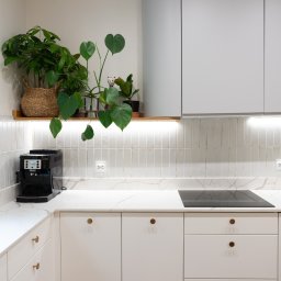 Triangle Studio - Jasna kuchnia z białymi szafkami, blatem imitującym marmur i roślinami doniczkowymi na półce. Ekspres do kawy na blacie. Minimalistyczny design z akcentami drewna.