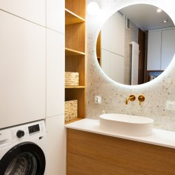 Triangle Studio - Nowoczesna łazienka z okrągłym lustrem podświetlanym LED, umywalką nablatową, złotą armaturą i pralką. Jasne drewno szafek i terazzo na ścianie.