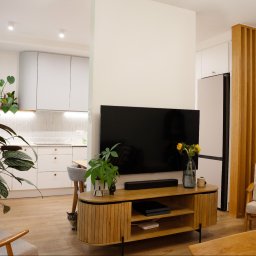 Triangle Studio - Jasne wnętrze mieszkania z otwartą kuchnią, drewnianą podłogą i meblami w stylu skandynawskim. Widoczne rośliny doniczkowe i kot.