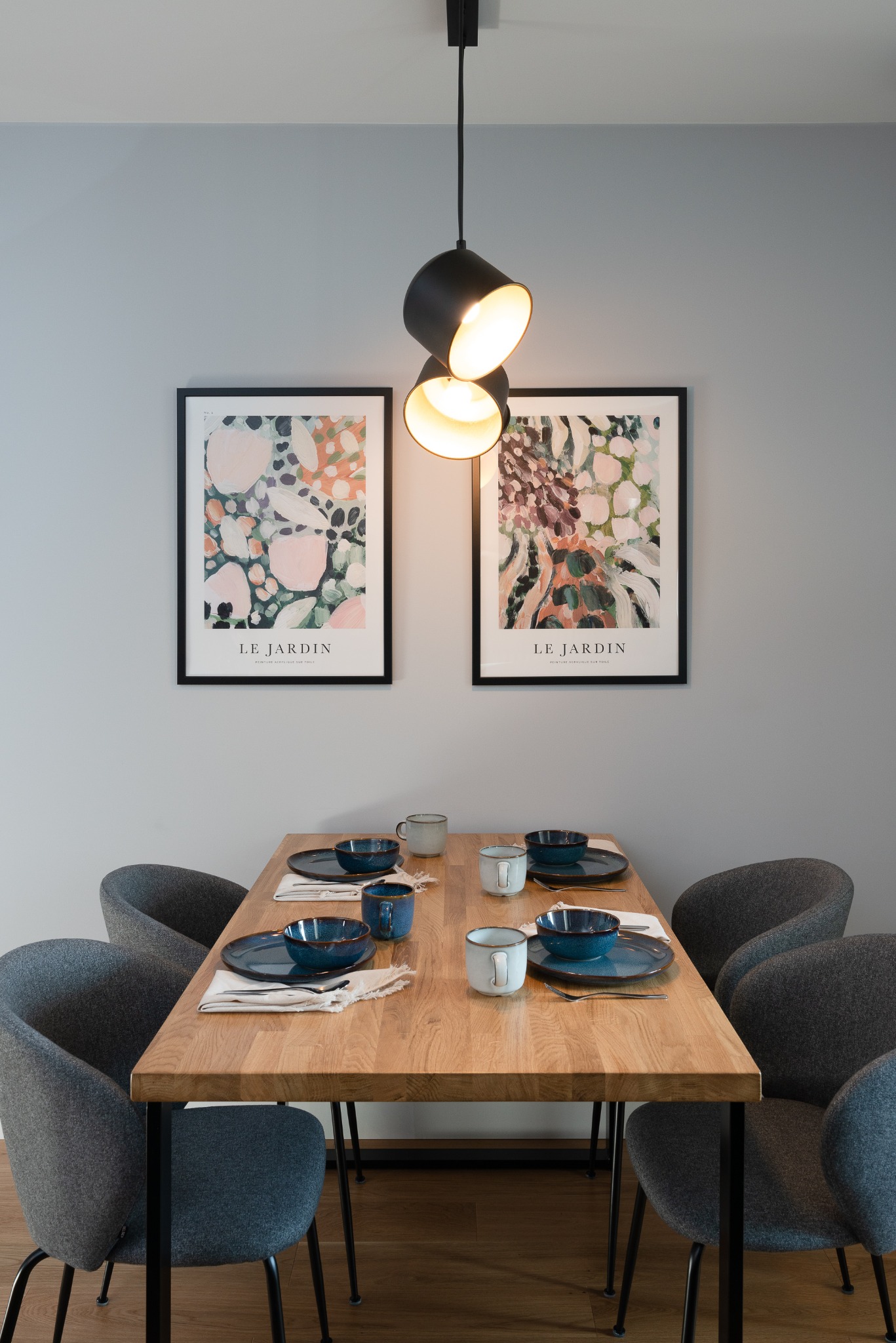 Stół z drewna dębowego, nakryty dla czterech osób, z niebieską zastawą i szarymi krzesłami. Nad nim designerska, czarna lampa. Na ścianie grafiki w czarnych ramach.