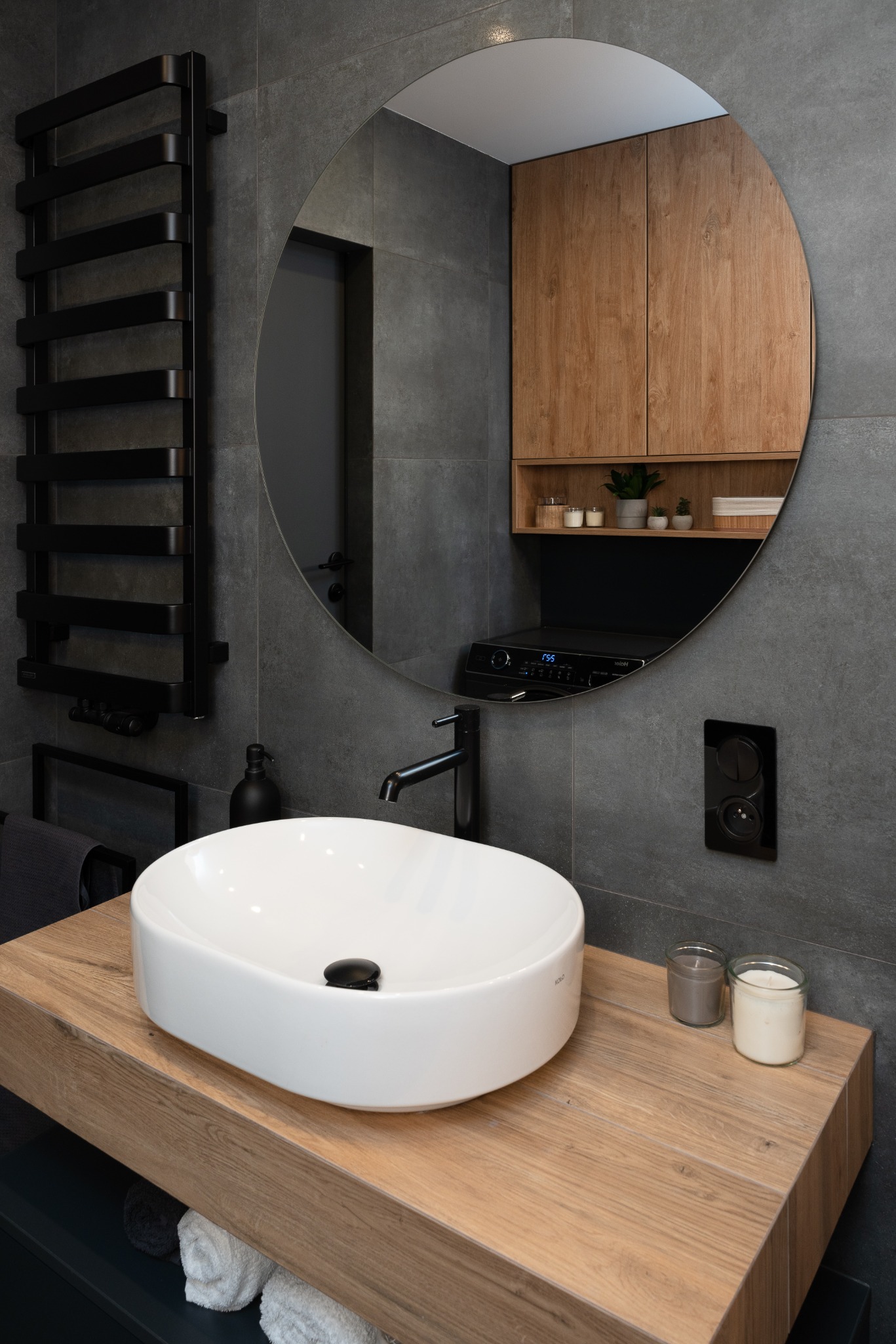 Nowoczesna łazienka z owalną umywalką nablatową KOLO na drewnianym blacie, czarną armaturą i grzejnikiem drabinkowym na tle szarych płytek.