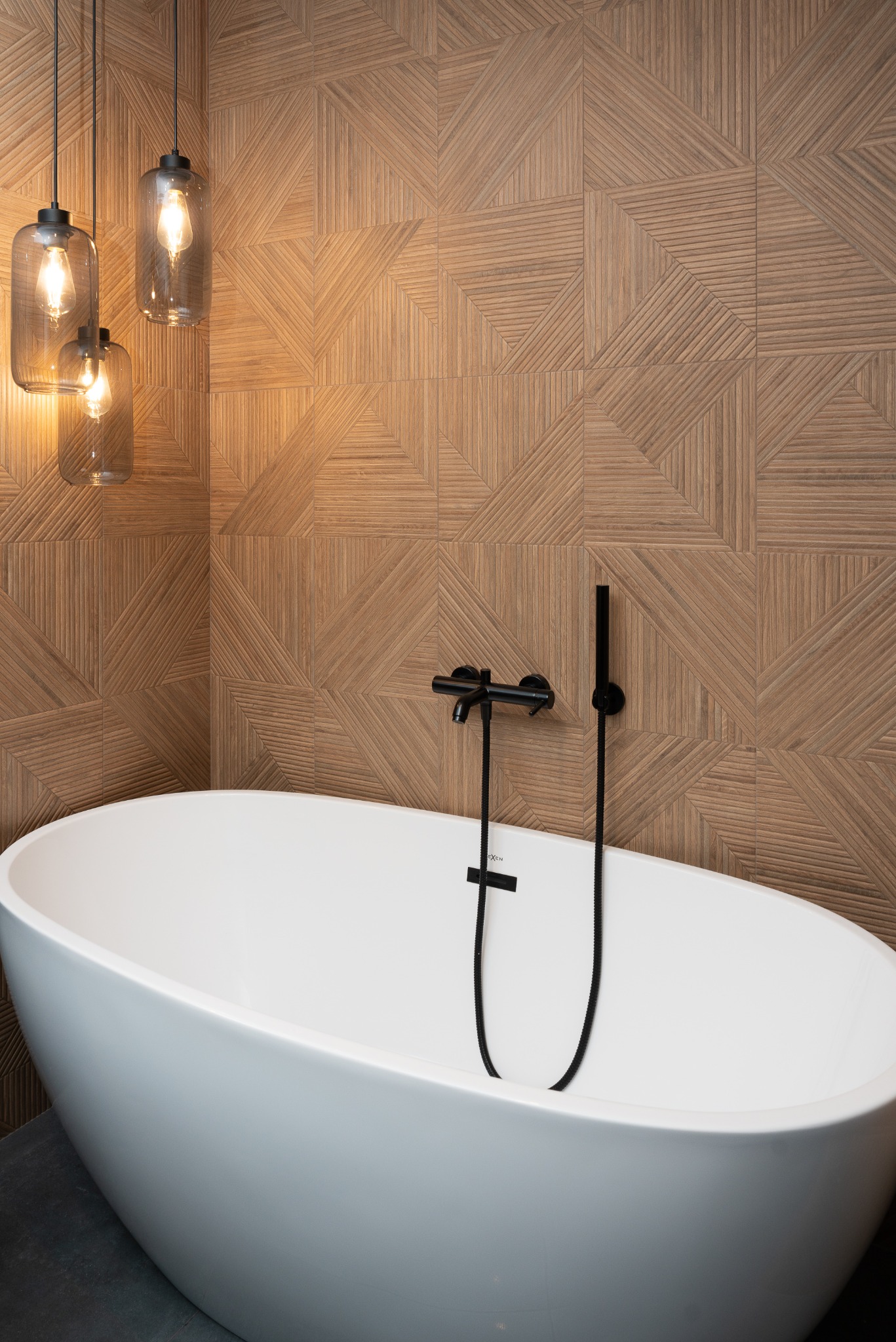 Nowoczesna łazienka: wolnostojąca wanna, geometryczna ściana z drewnopodobnych płytek, czarna armatura i wiszące lampy z żarówkami Edisona.