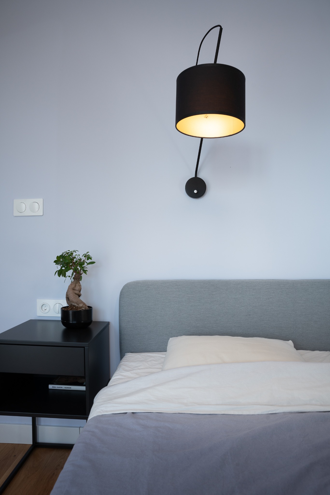 Minimalistyczna sypialnia: szare łóżko z zagłówkiem, czarna szafka nocna z bonsai, designerska lampa ścienna na tle jasnoniebieskiej ściany z włącznikami.