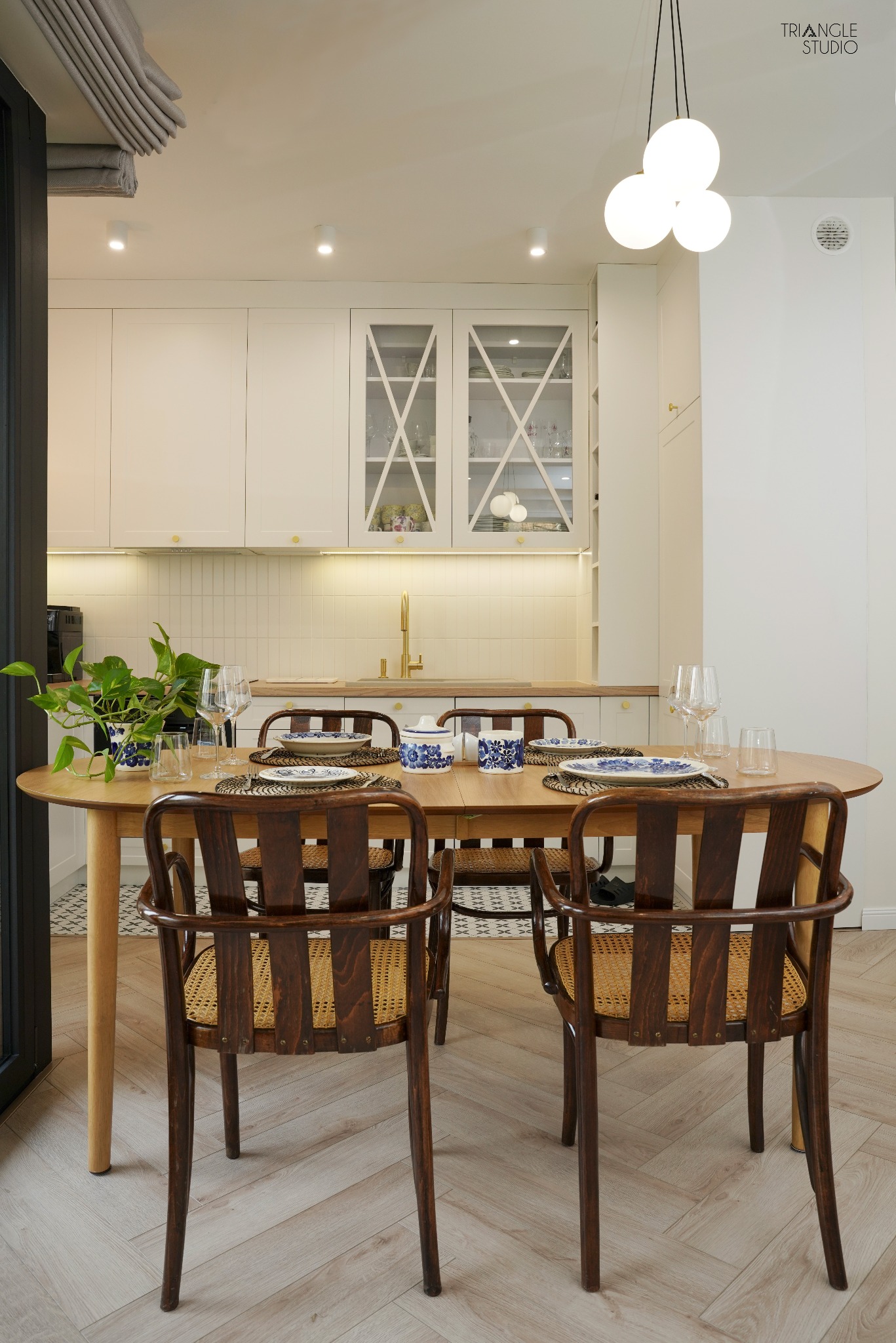 Jasna kuchnia z drewnianym stołem i krzesłami w stylu retro. Na stole zastawa w niebieskie wzory. W tle białe szafki z przeszklonymi frontami i złota armatura.