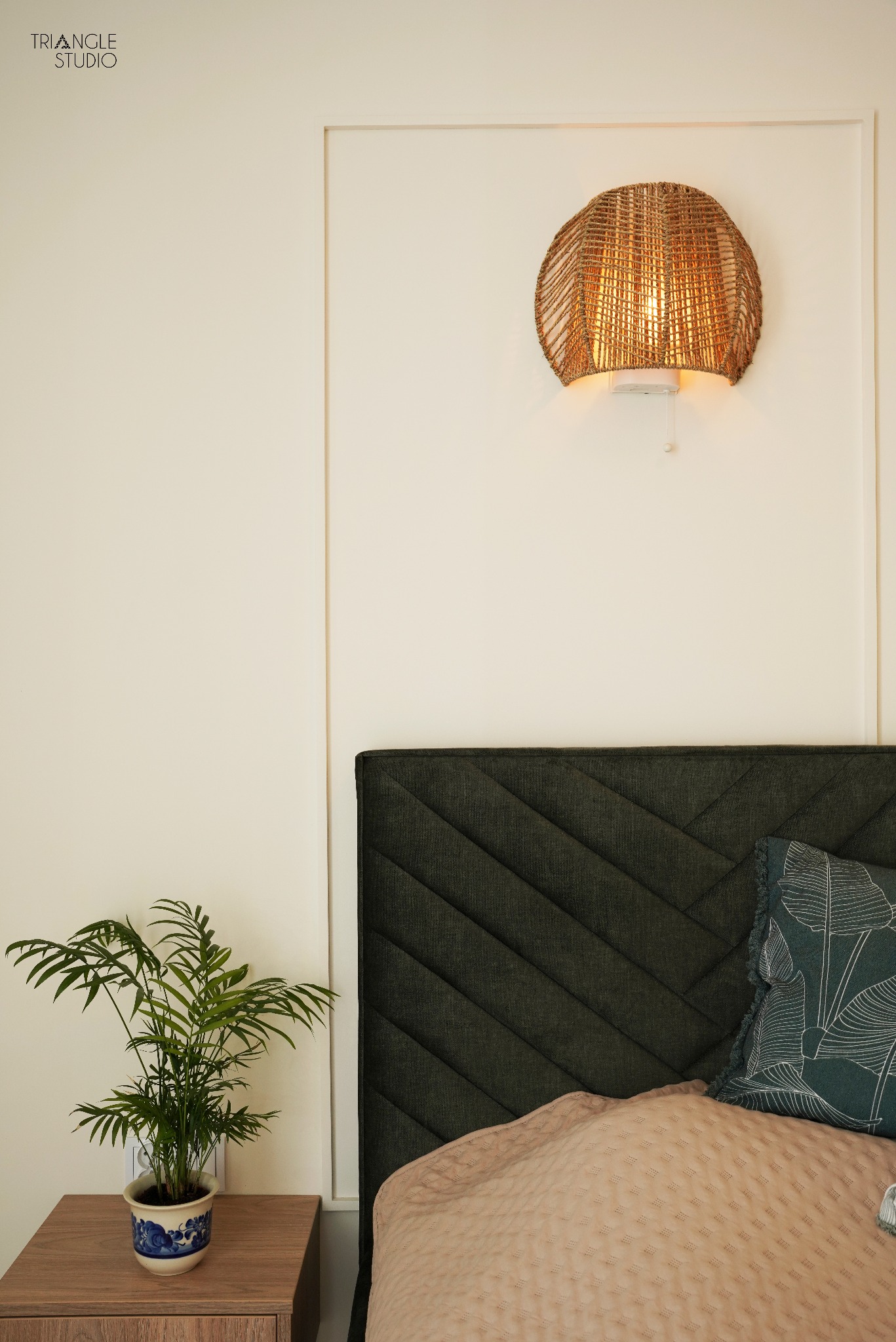 Fragment sypialni: wezgłowie z zielonej tkaniny w jodełkę, pleciony abażur lampy na ścianie, roślina doniczkowa na szafce nocnej. Minimalistyczny design, ciepłe oświetlenie.