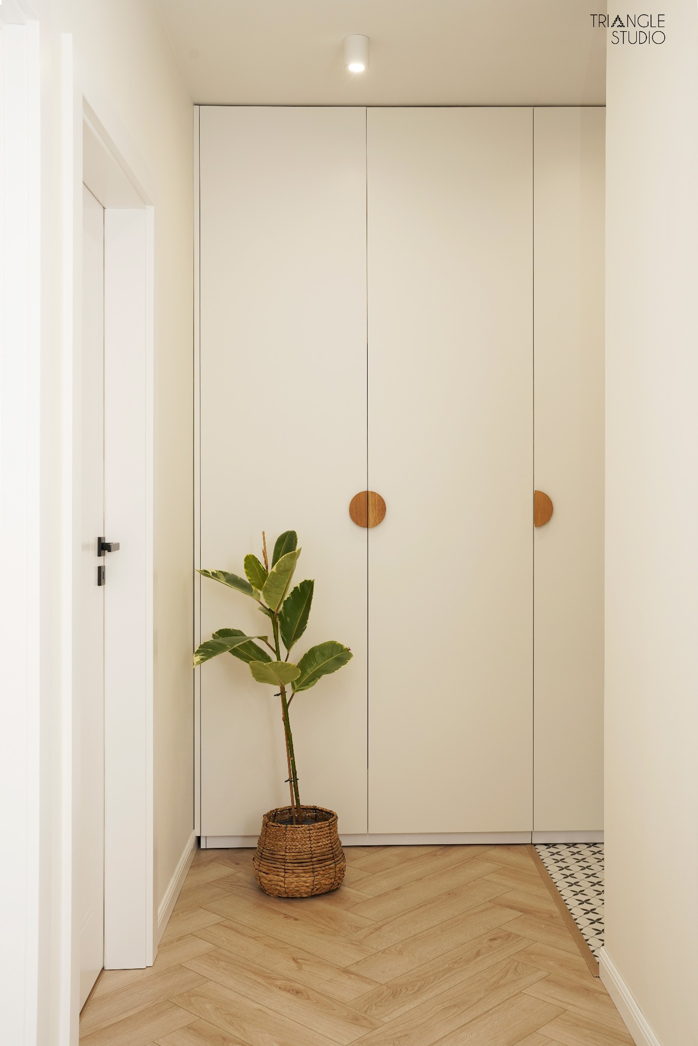 Minimalistyczny przedpokój z parkietem w jodełkę, białą zabudową szaf z okrągłymi drewnianymi uchwytami i rośliną w wiklinowym koszu. Widoczny fragment wzorzystej podłogi.